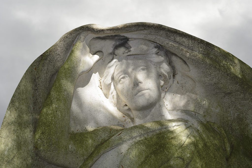 Photograph of an angel sculpture from chest up, with wings making a large rounded hood covering. There are hints of sunlight across her face. The sky is grey and clouded.
