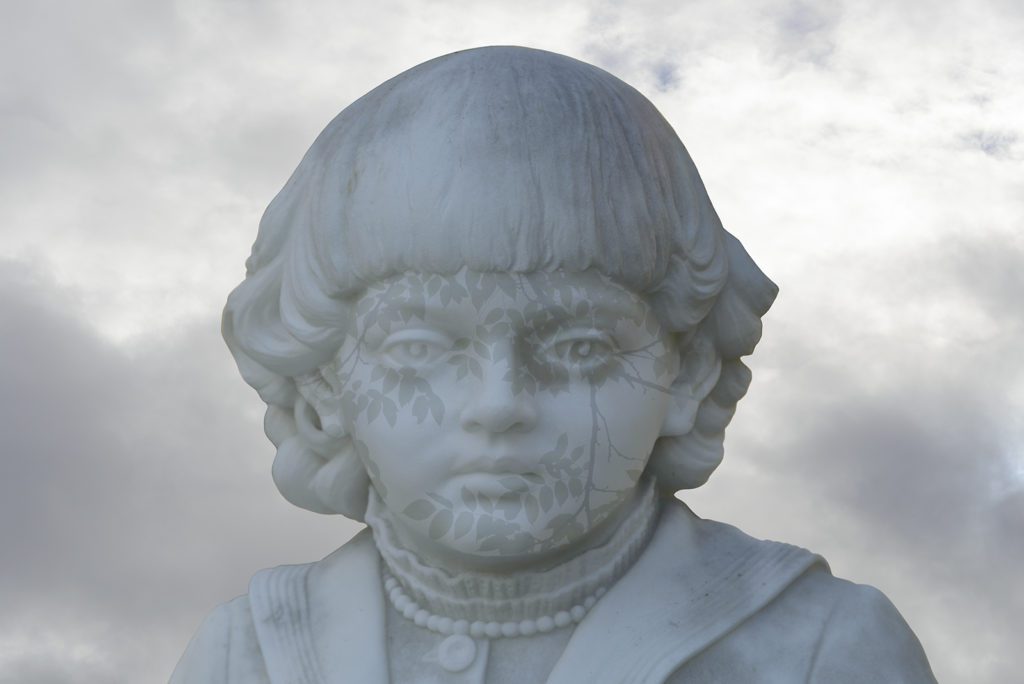 Photograph of a child in marble with a placid expression, short wavy hair, and a high collard shirt with beaded necklace. A shadow leaf pattern has been imposed over her face. The sky is grey and clouded.