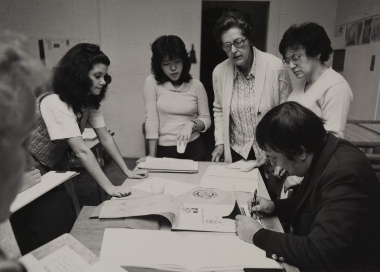 1970s black and white photograph of adult students standing around a table. On the far right is a man, seated at the table holding a drawing demonstration.