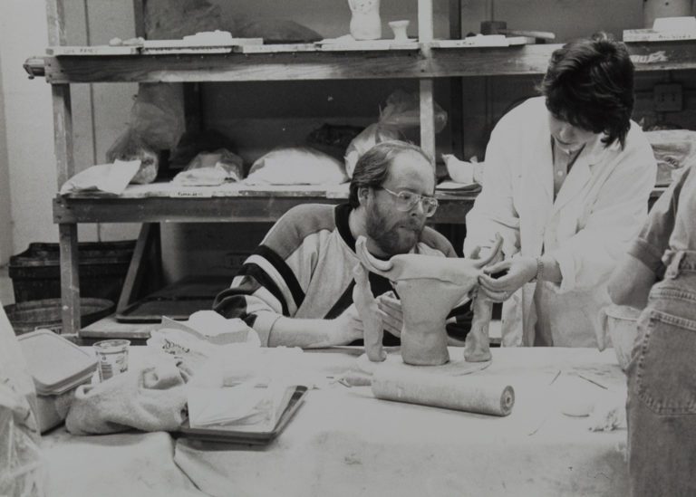 1970s photograph of a man, kneeling at a table with sculpting materials on it, and a woman in a long white smock handling a sculpture piece.