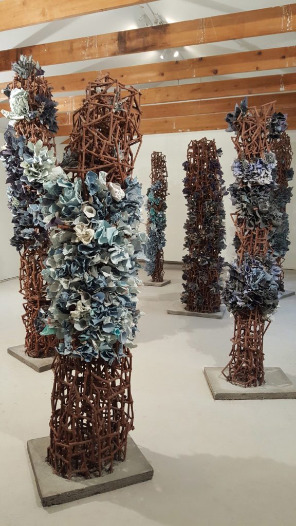 Photograph of tall skinny sculptures on grey flat square bases. Each has rusted scaffolding with sections of blue, grey, and white paper stuffed into it making floral tubes.