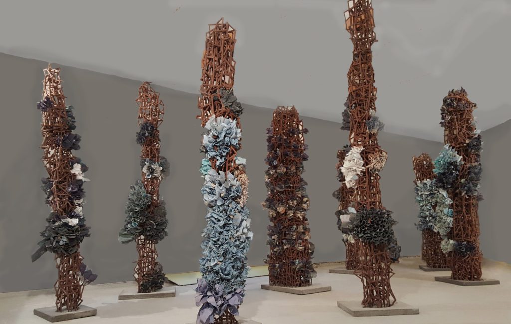Photograph of tall skinny sculptures on grey flat square bases. Each has rusted scaffolding with sections of blue, grey, and white paper stuffed into it making floral tubes.
