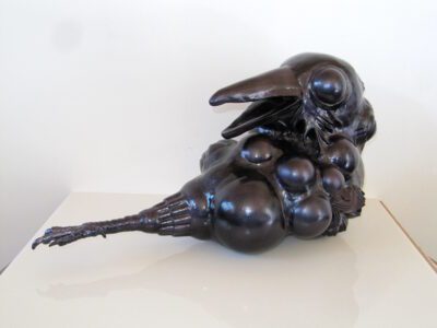 Metal sculpture in black of a raven's head looking back over its body. Its body is made of circular globs, has no feet or wings, but a long tail like structure that ends in a single foot.