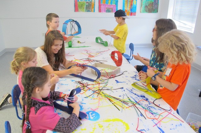 Photograph of a group of young students at a long white table making sculptures out of paper and string.