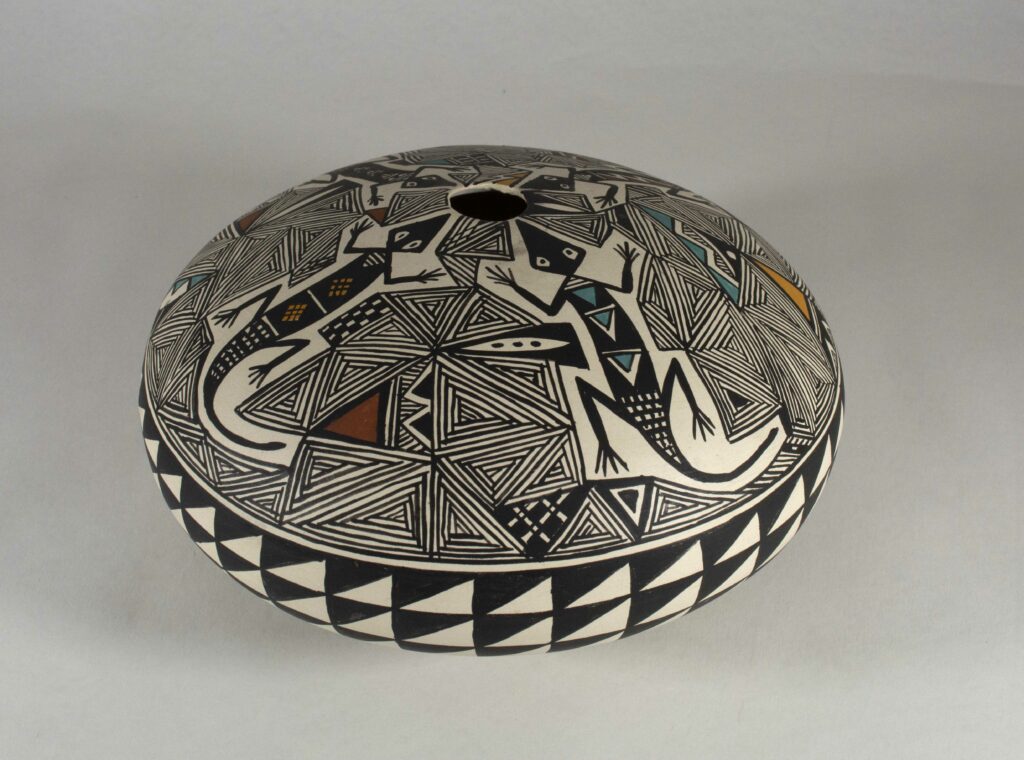 White and black flat bowl with a very small opening in the top center. There are triangular patterns painted all over with hints of colors of red, teal, and yellow. There are also four lizards radiating from the center hole.