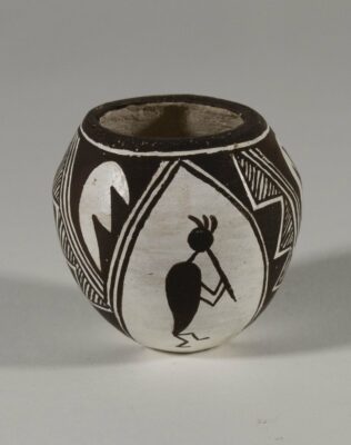 Small black and white pot with geometric designs painted on the front. In the center is a dancing figure in profile playing a long flute.