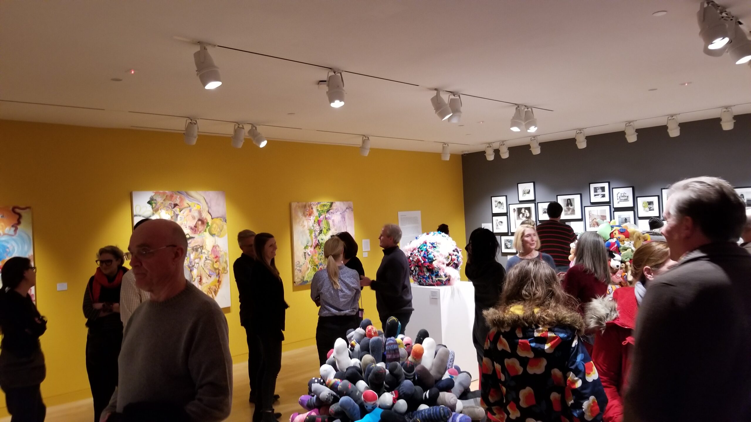 Photograph of a gallery room with yellow and grey walls. There are many people standing about talking and looking at the artwork.