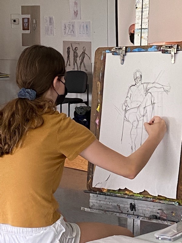 Photograph of a teenaged girl in a mustard orange t-shirt seated at an easel drawing a model.