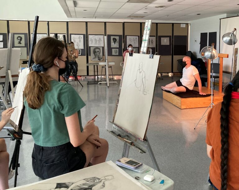 Photograph of a teenaged girl in a green t-shirt seated at an easel drawing a model.