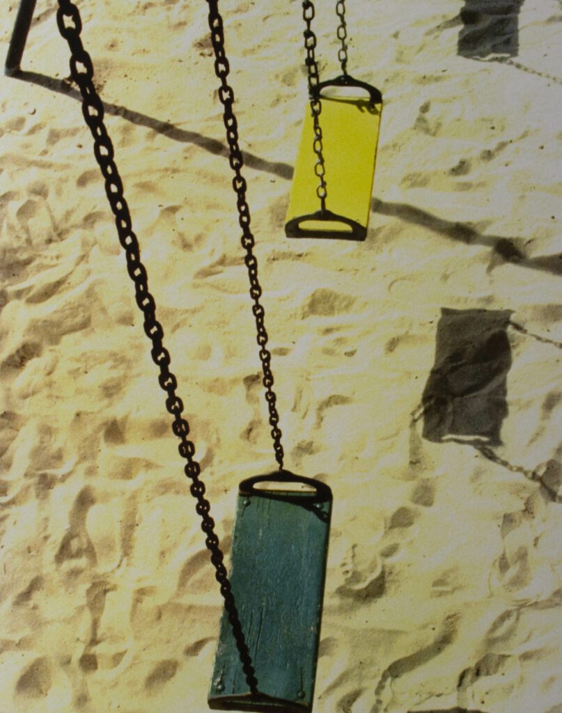 Yellow saturated photograph of a sandy playground with two wooden swings hanging from chains. One is painted blue and the other yellow.