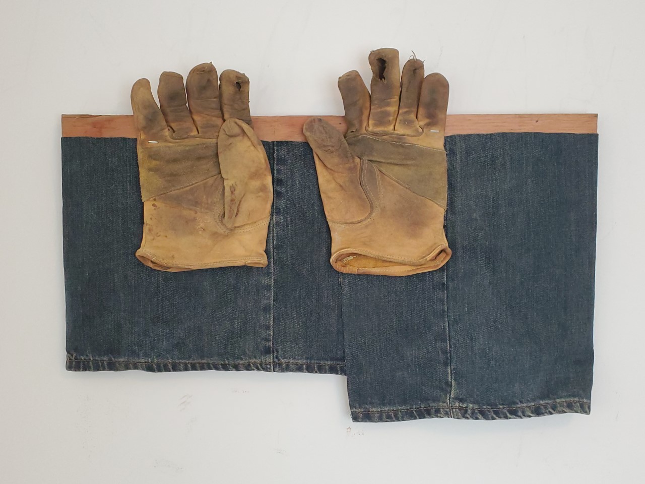 Photograph of two pieces of jean fabric hanging from a wooden bar. There are two yellow work gloves stapled to the bar.