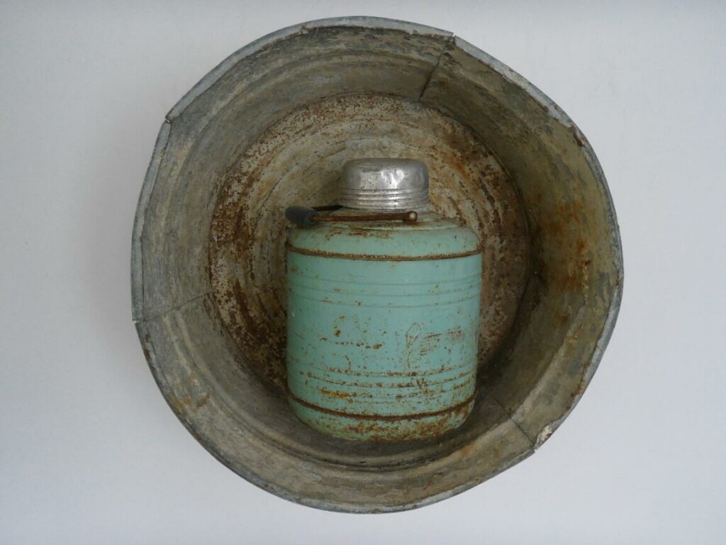 Photograph of the inside of a rusted grey bucket holding a teal canteen with a handle and silver top.