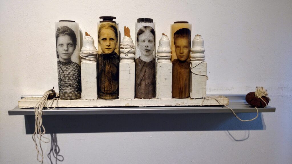 Sculpture resting on a metal platform hanging on the wall. There are white railings cut towards the bottom with old paint rollers between the railings, wrapped in plastic with old photographs of girls printed on them.