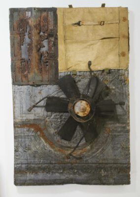 Mixed media wall hanging with rusted tin plates in the background, a piece of rusted vent grate on the upper left, canvas on the upper right, and an industrial fan blade in the center.