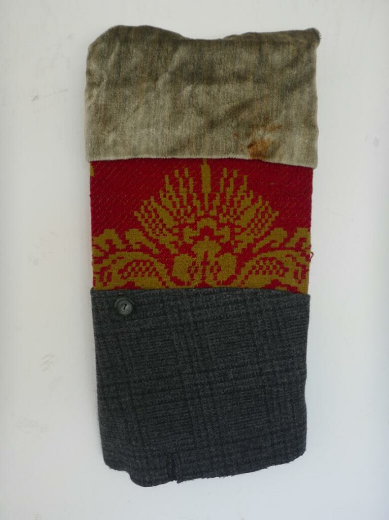 Photograph of a fabric rectangle with a plaid dark grey fabric, red center fabric with gold detailing, and a silk gold-brown top.