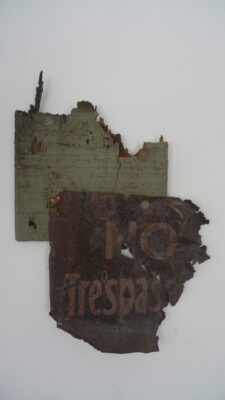 Photograph of two old pieces of signs, one rusted and one wood, nailed together. The metal sign reads 'No Trespass...'.