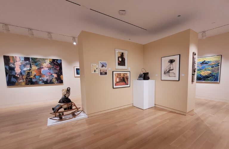 Photograph of a gallery room with tan walls and tan wood floor. There are various works hanging on the walls and a few founds sculptures on pedestals.