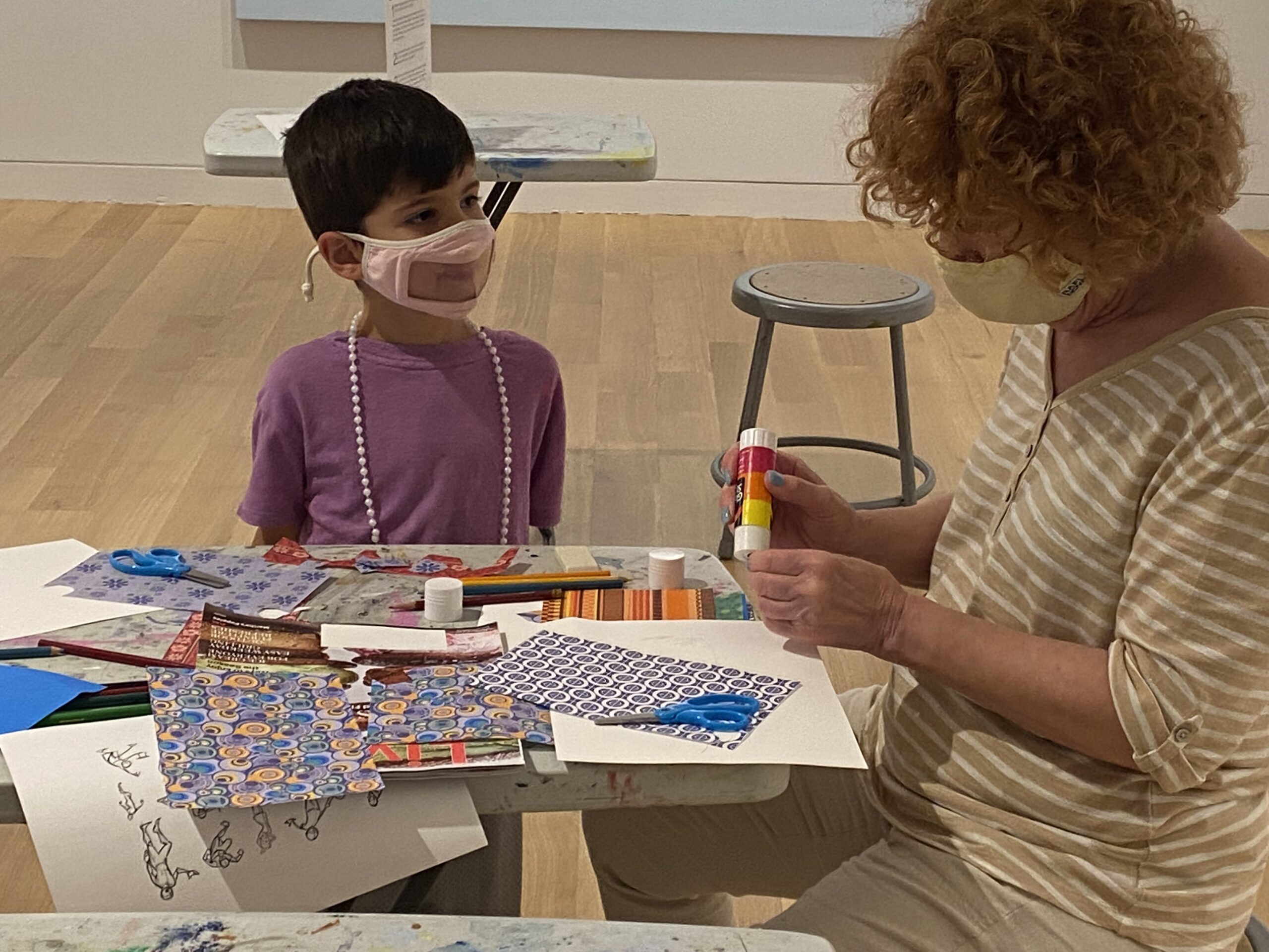 Photograph of a boy with short dark hair and pink t-shirt seated at a small table filled with colorful papers. An older woman is sitting on the right with short red curly hair gluing paper.