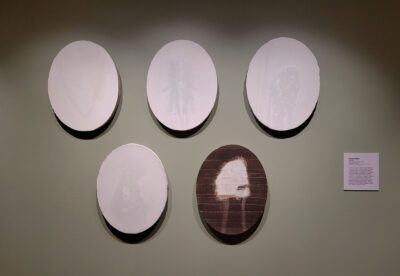 Photograph of five oval works on a green wall, Four works are white with ghostly images, the fifth is brown with a white center.