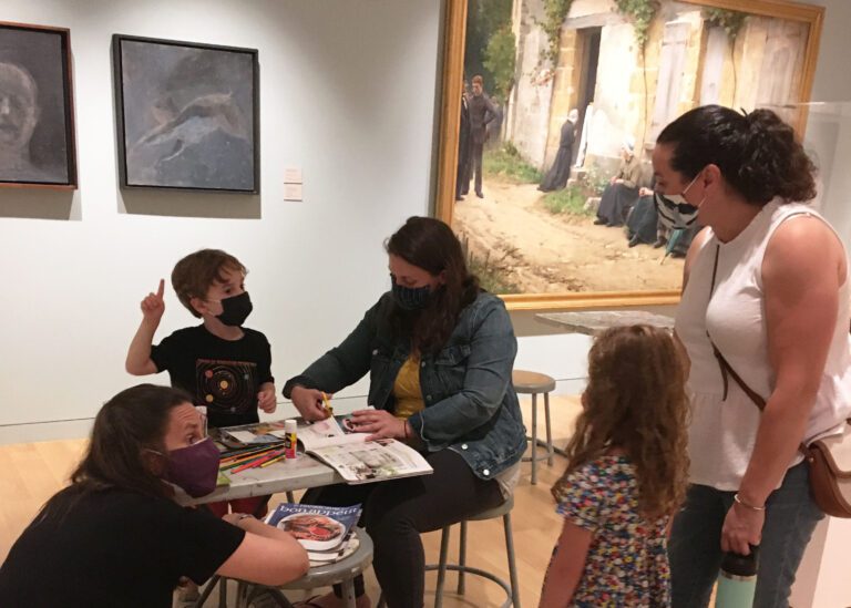 Photograph of two families of two in a museum gallery, standing around a table with a woman crouched near it cutting out images from a magazine.