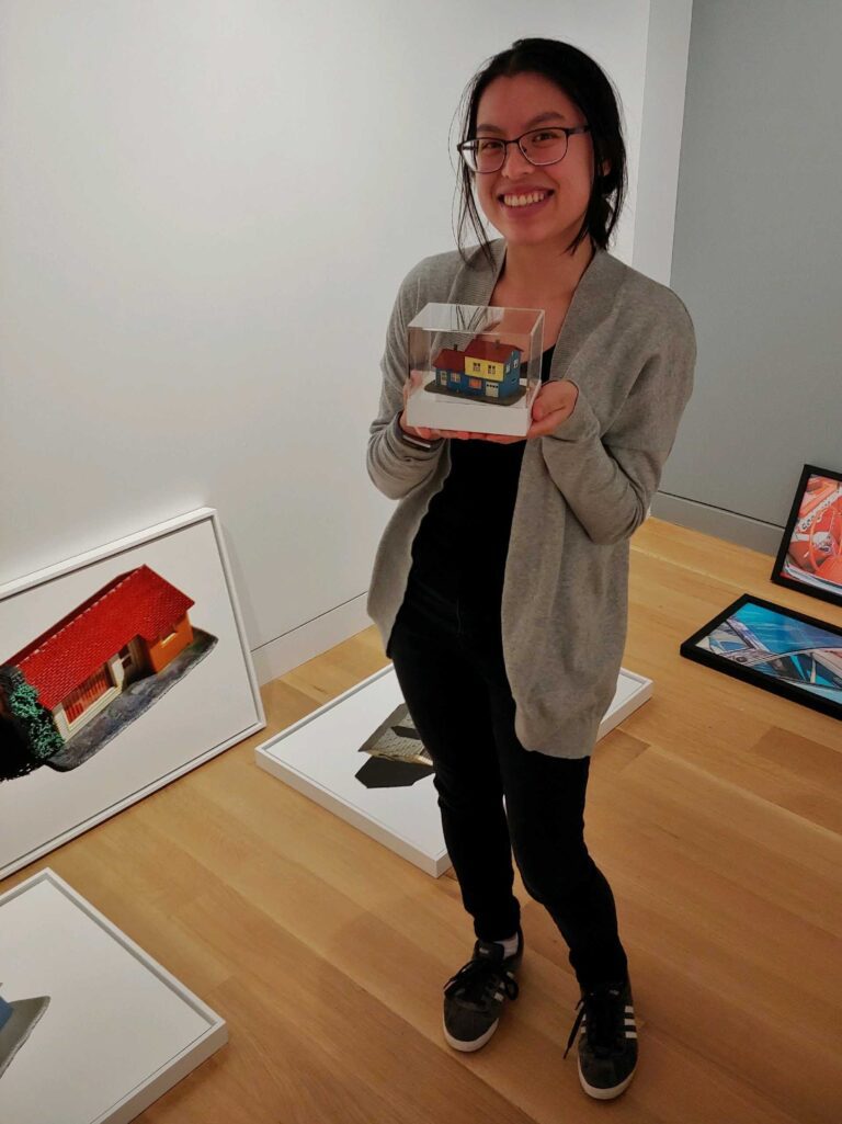 Photograph of a woman with long black hair, black outfit, and grey sweater. She is standing in a gallery room with paintings on the floor, holding a small model house in an acrylic box.
