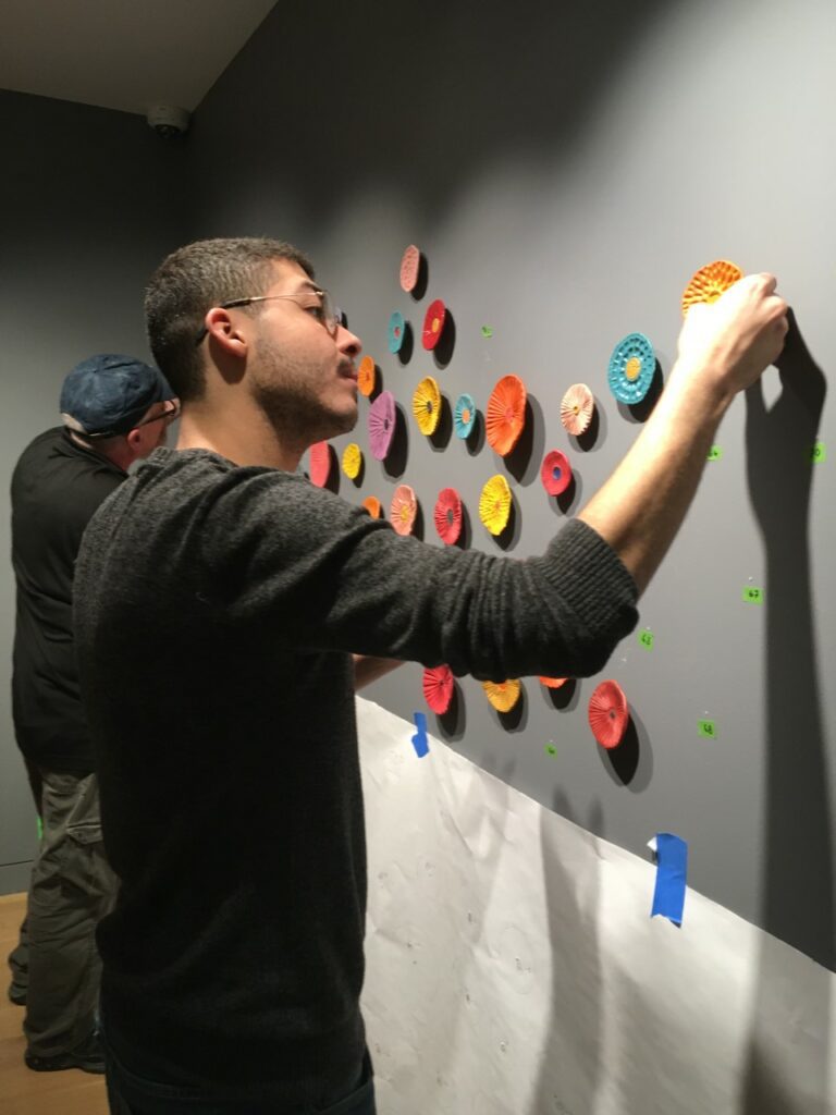 Photograph of a man with short brown hair and grey sweater, standing at a grey wall installing ceramic flowers.