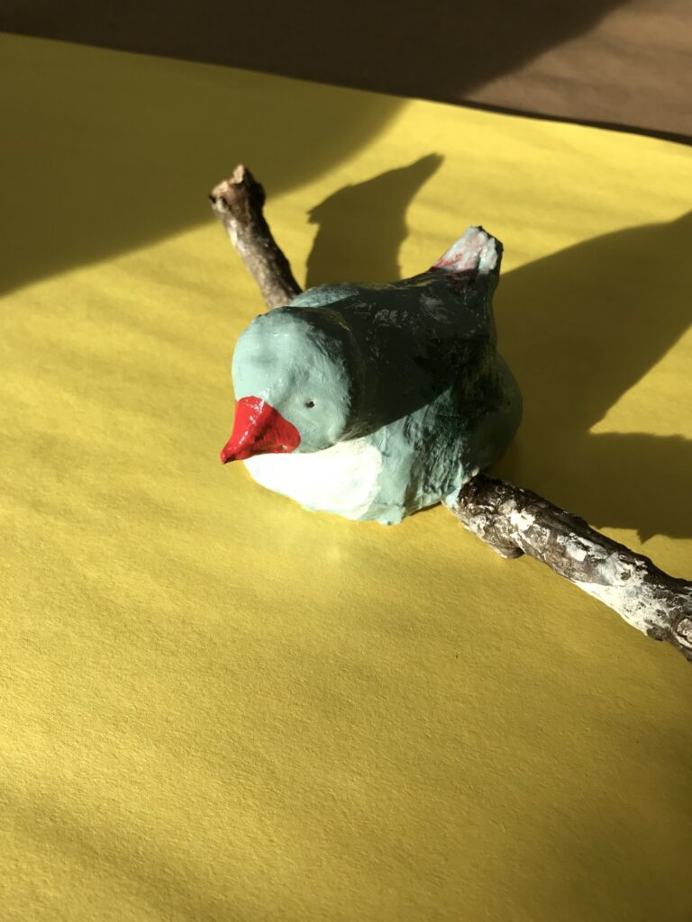 Photograph of a blue ceramic bird on a stick, taken from a yellow table surface with sunlight creating dramatic shadows behind it.