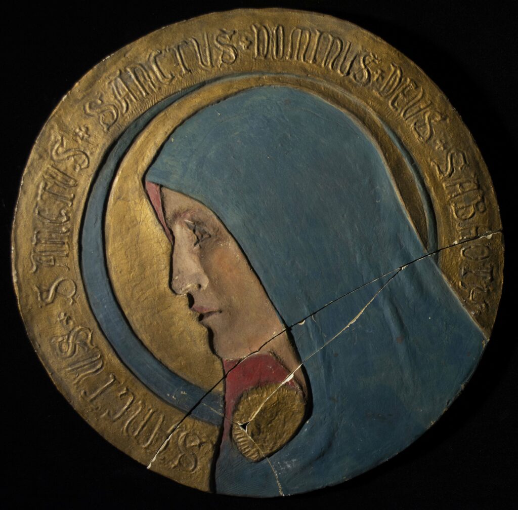 Round plaque with a gold border surrounding a woman in profile from the shoulder up. She is wearing a blue hooded garment with red interior, and she has a halo behind her.
