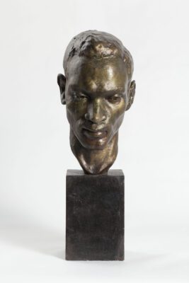 Portrait bust on a tall rectangular base of an African man, looking slightly downward. His face is narrow and angular, with short cropped hair.