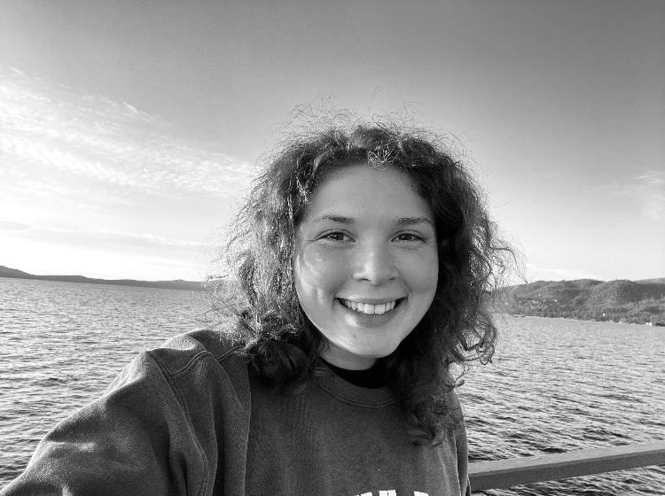 Black and white photograph of a young white woman with fuzzy hair in a sweat shirt in front of a body of water.