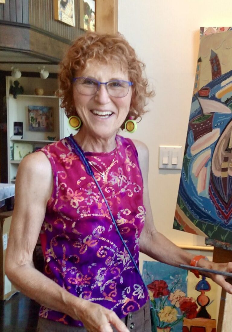 Photograph of a white woman with short curly red hair, in a bright pink and purple top. She is standing at an easel with an abstract painting, more of which are hanging in the background.