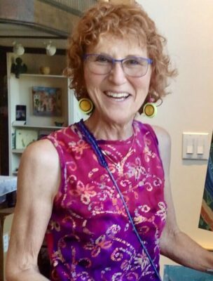 Photograph of a white woman with short curly red hair, in a bright pink and purple top. She is standing at an easel with an abstract painting, more of which are hanging in the background.