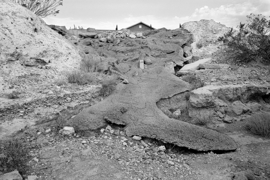 Black and white photograph of rocky outdoor scene with a flow of magma like rock down the middle from the sloping up ward hill. Behind the hill just visible is the peak of a house roof.