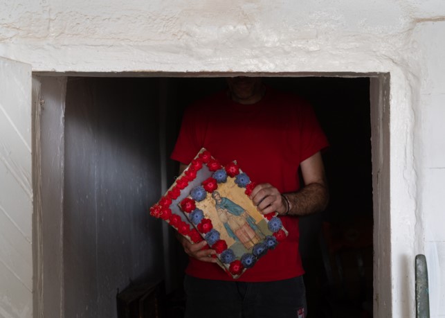 Photograph of half of a small door surrounded by white stucco. In the doorway is visible the torso of a man in a red shirt holding two rectangles, one with blue and red flowers around the perimeter, with a painted saint in the center.