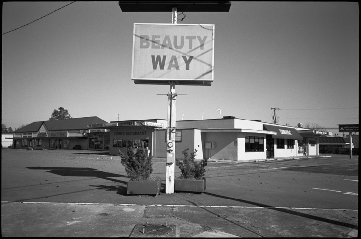 Black and white photograph of a strip mall in a paved parking lot. In the center is a sign that states 'Beauty Way'.
