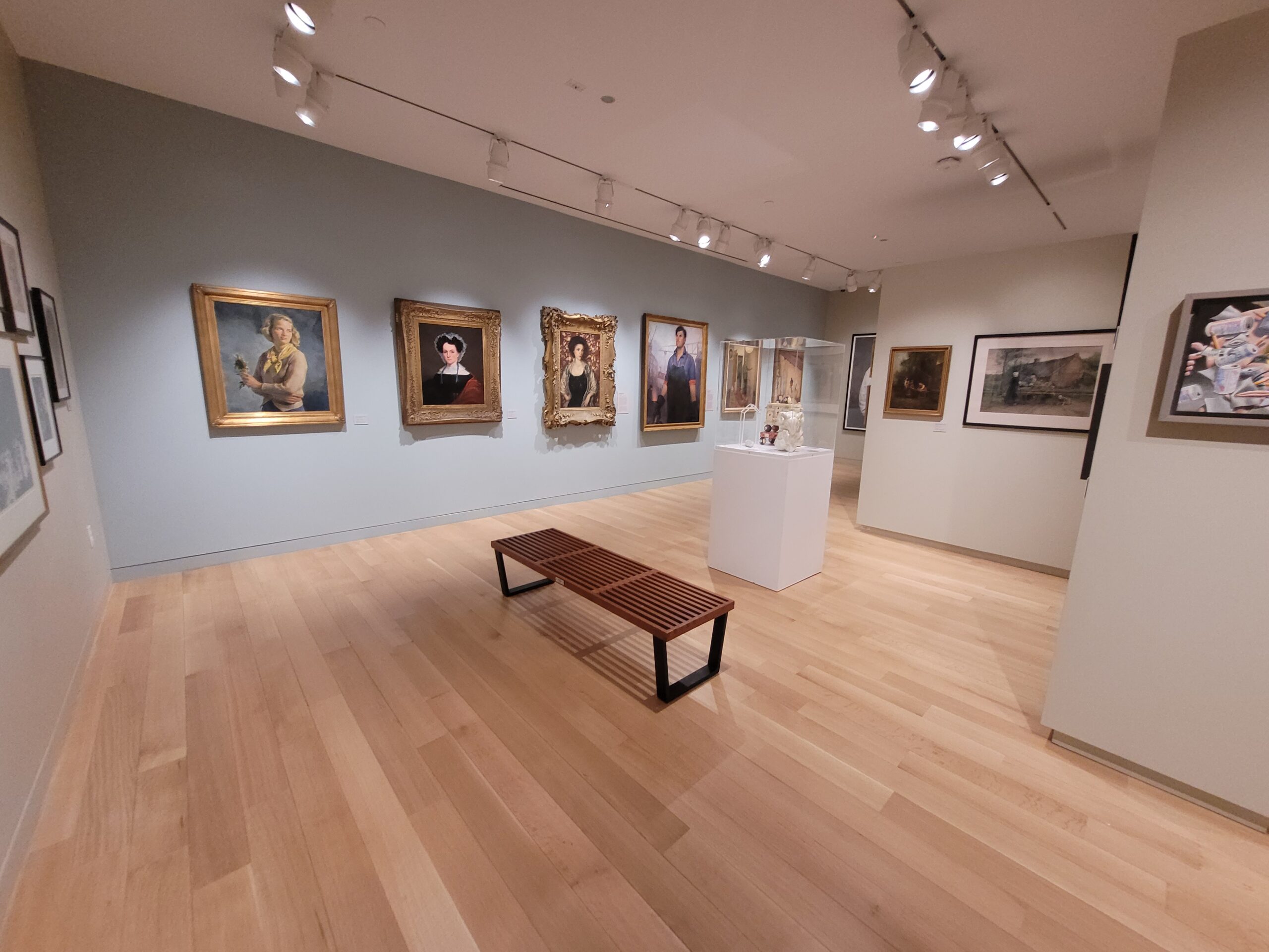 Photograph of a gallery installation with blue and grey-green walls and light brown wood floors. There are various artworks hanging on all the walls, and a floor case with clear top is holding three sculpture works.