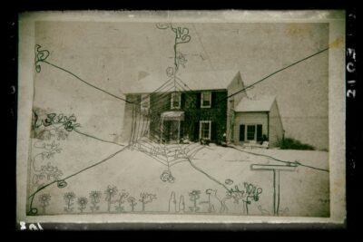 Black and white photograph of a two story house with a brick façade in the winter. Drawn on top is a spider web in the center with lines coming out to the edges of the photograph, and flowers, bottles, and a cat along the bottom.