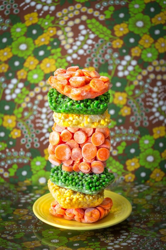 Photograph with a colorful floral background in greens, yellow, white, and blue. In the foreground is a yellow plate with stacks of frozen veggies of carrots, corn, and peas.