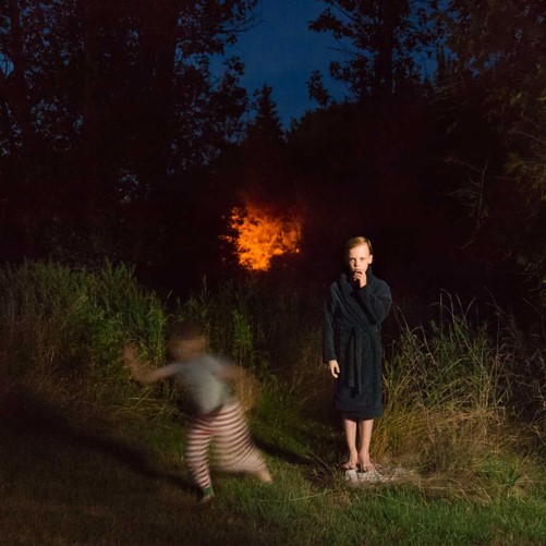 Photograph of a night scene with a boy standing in a bathrobe with a hand up to his chin and a second boy with stripped pants blurred running to the left. In the background are the outline of trees, but in the center is a bright red spot almost like a fire.
