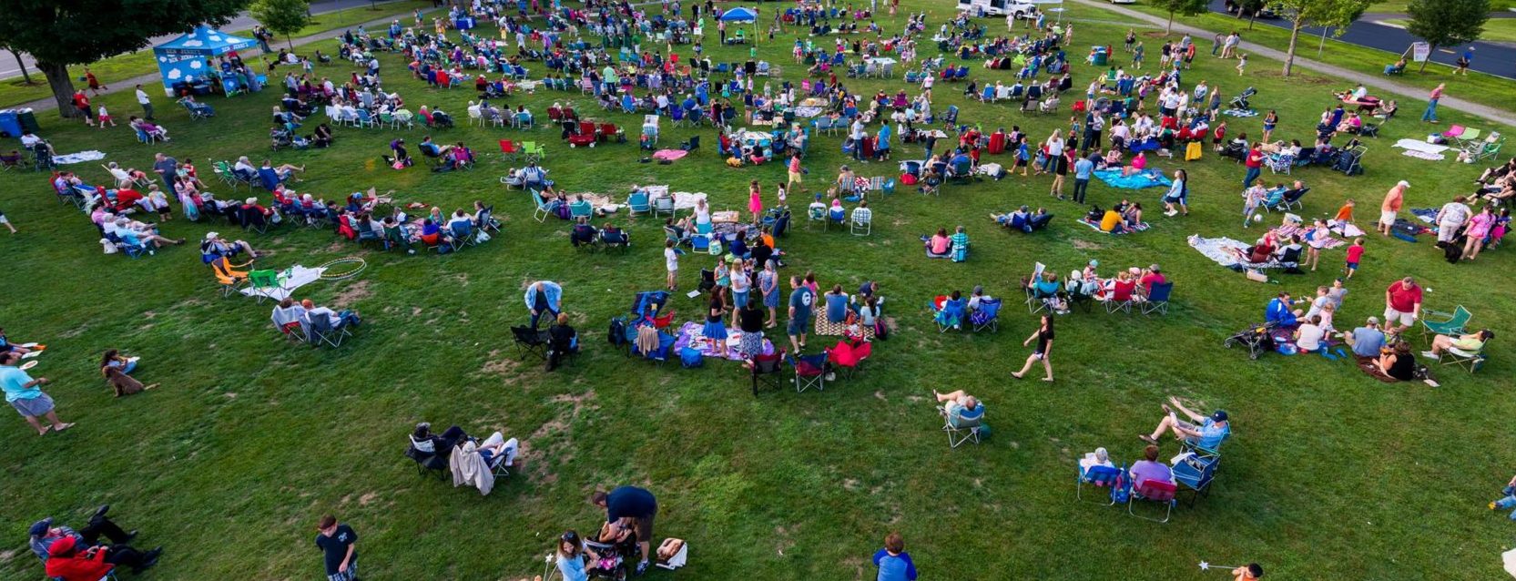 Wide angled areal photograph of an outdoor lawn with many people seated on the lawn.