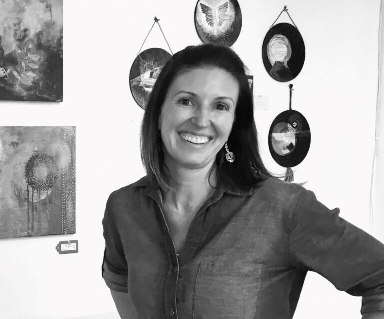 Black and white photograph of a woman with short straight dark hair and dangly earrings. She is looking at the camera with one arm on her him, standing in front of artwork and bowls hanging on the wall behind her.