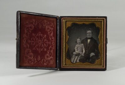 Photograph of an old glass negative in a leather and felt case. The photograph is of an older man in a 19th century suit with a young girl by his side in a dress with ringlet hair.