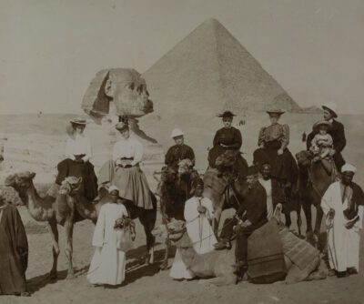 Late 19th century photograph of a tour group in Egypt with the pyramids and sphynx in the background. The group is mostly women, with two men and one child, all on camels. Tour guides in white robes are standing in front of the group.