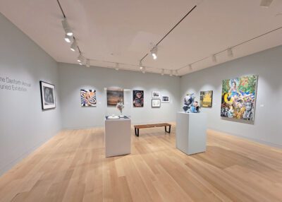 Photograph of a gallery with grey walls and light wood floor. There are various paintings hung on the walls, and two pedestals containing sculpture pieces in the center along with a bench.