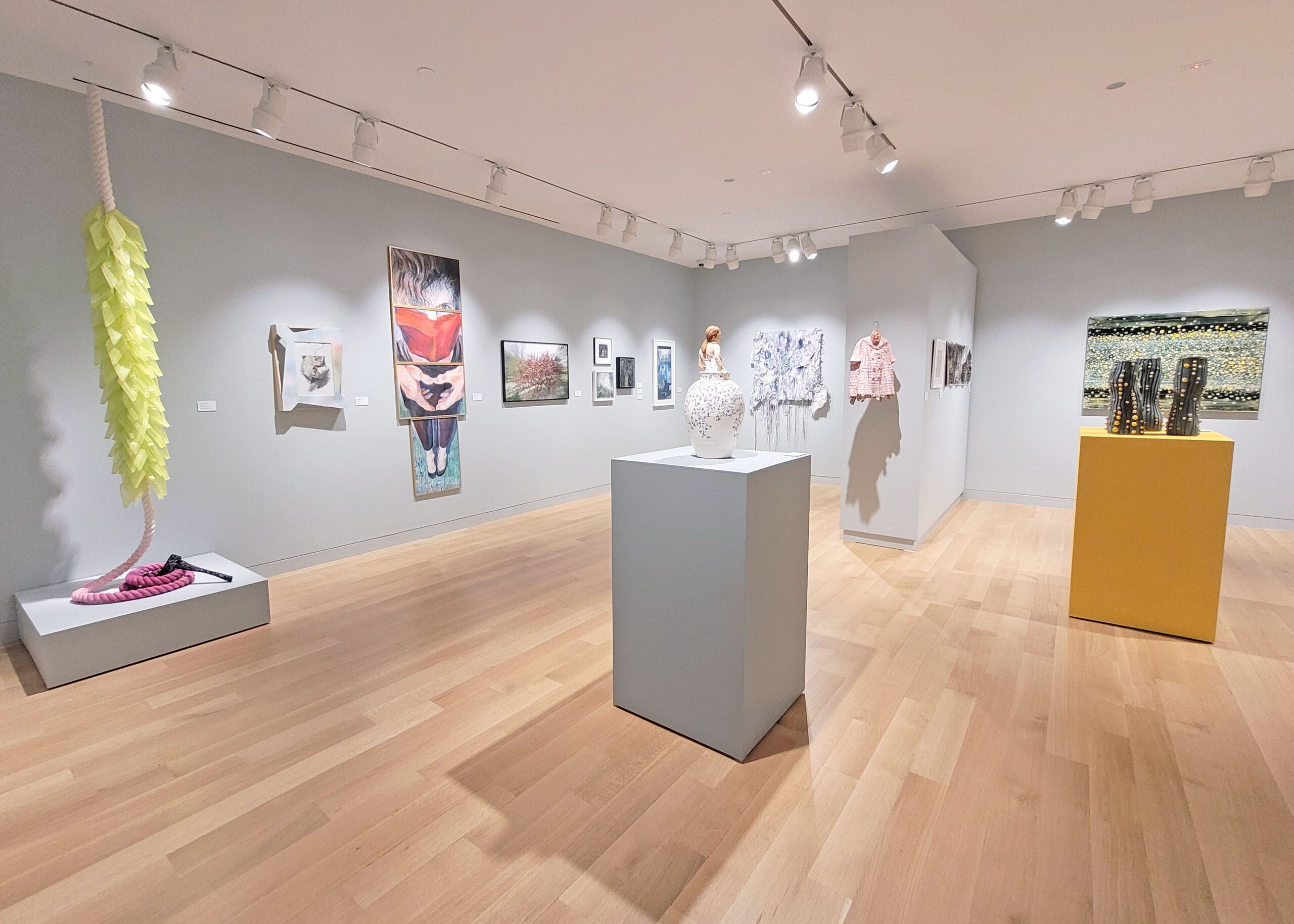 Photograph of a gallery installation with grey walls. There are various works hung on the walls, including two fabric pieces hanging from the ceiling. There are also to pedestals, one grey and one yellow, with ceramic works on top.
