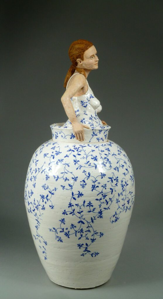 Photograph of a white vase with blue vine details. The top of the vase turns into the torso and head of a woman with a dress matching the vase. Her arms are out to the sides holding the vase rim, and her red hair is pulled back and down her back.