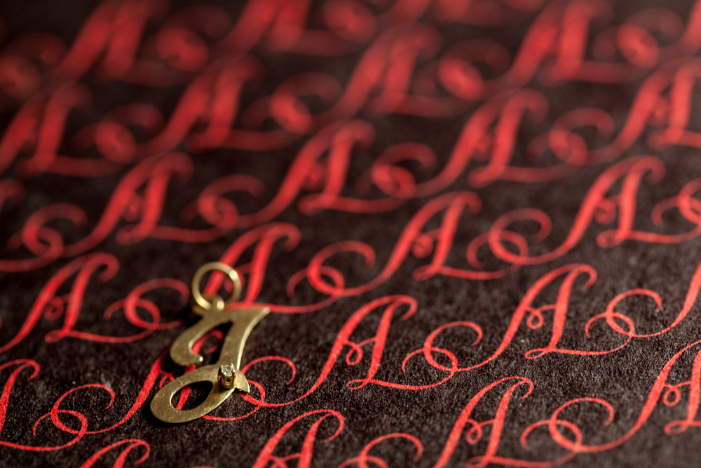 Photograph of a deep grey fabric with cursive capital 'A's across in a bright red. To the bottom left is a charm of a capital letter 'J' with a diamond towards the bottom of the 'J'.