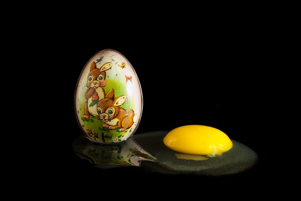 Photograph with a deep black surface and background. On the flat surface is an egg shaped sculpture with two drawn bunnies with a green background and butterflies around. To the right of the egg is an egg yolk with the whites surrounding, reflecting the sculpture egg.
