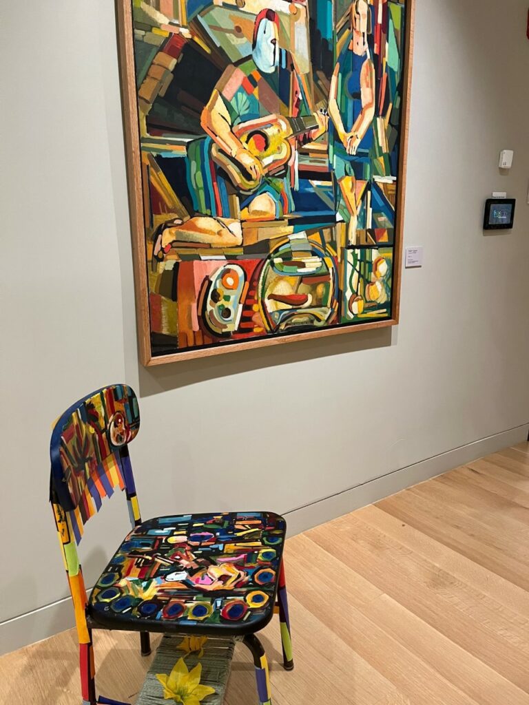 Photograph of an abstract drawing filled with lines and circles depicting a man and woman inside a room. In front of the painting is a decorated chair with various fabric, metal, and flowers finding inspiration from the painting behind it.