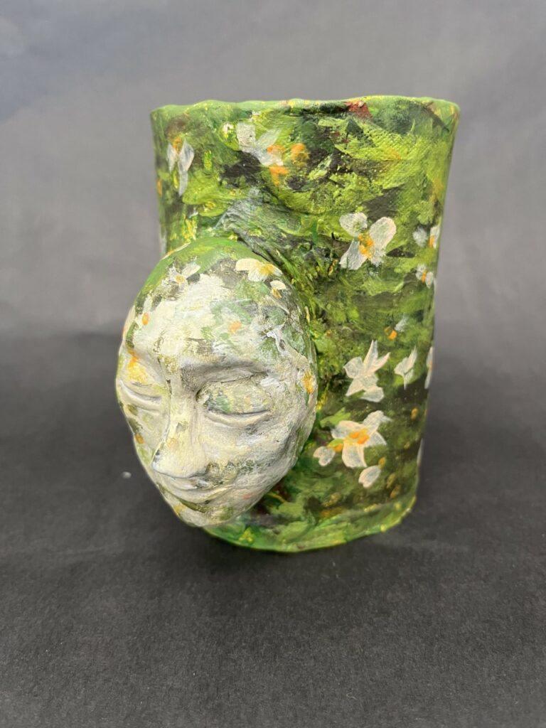 Photograph of a ceramic cup painted in green leaves and white flowers. On the side is a cream painted face with closed eyes.